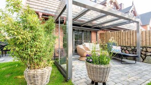 A covered patio area with wicker planters containing plants, a glass enclosure, and outdoor furniture.