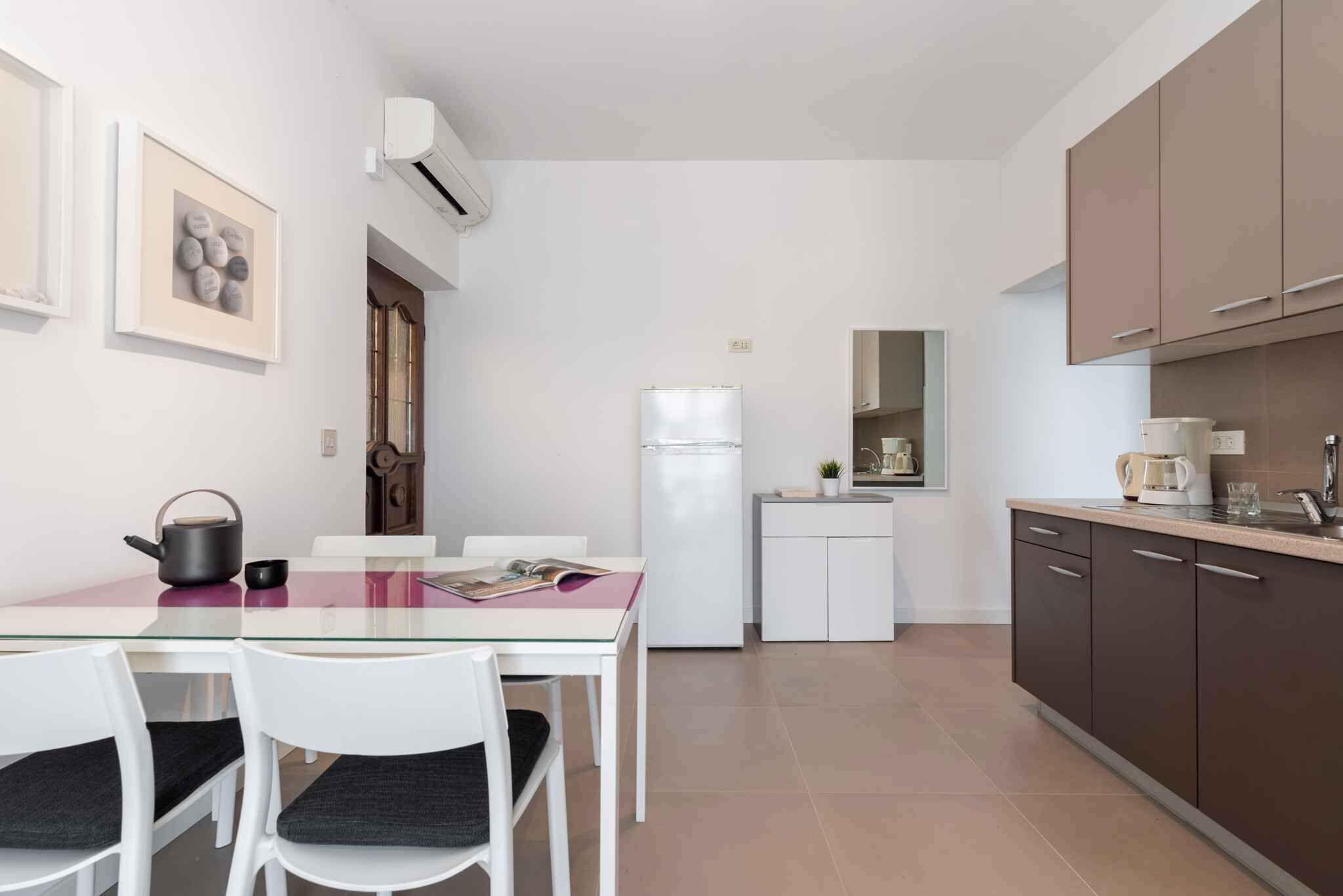 A compact kitchen with a glass dining table, white chairs, a white refrigerator, and brown cabinets.