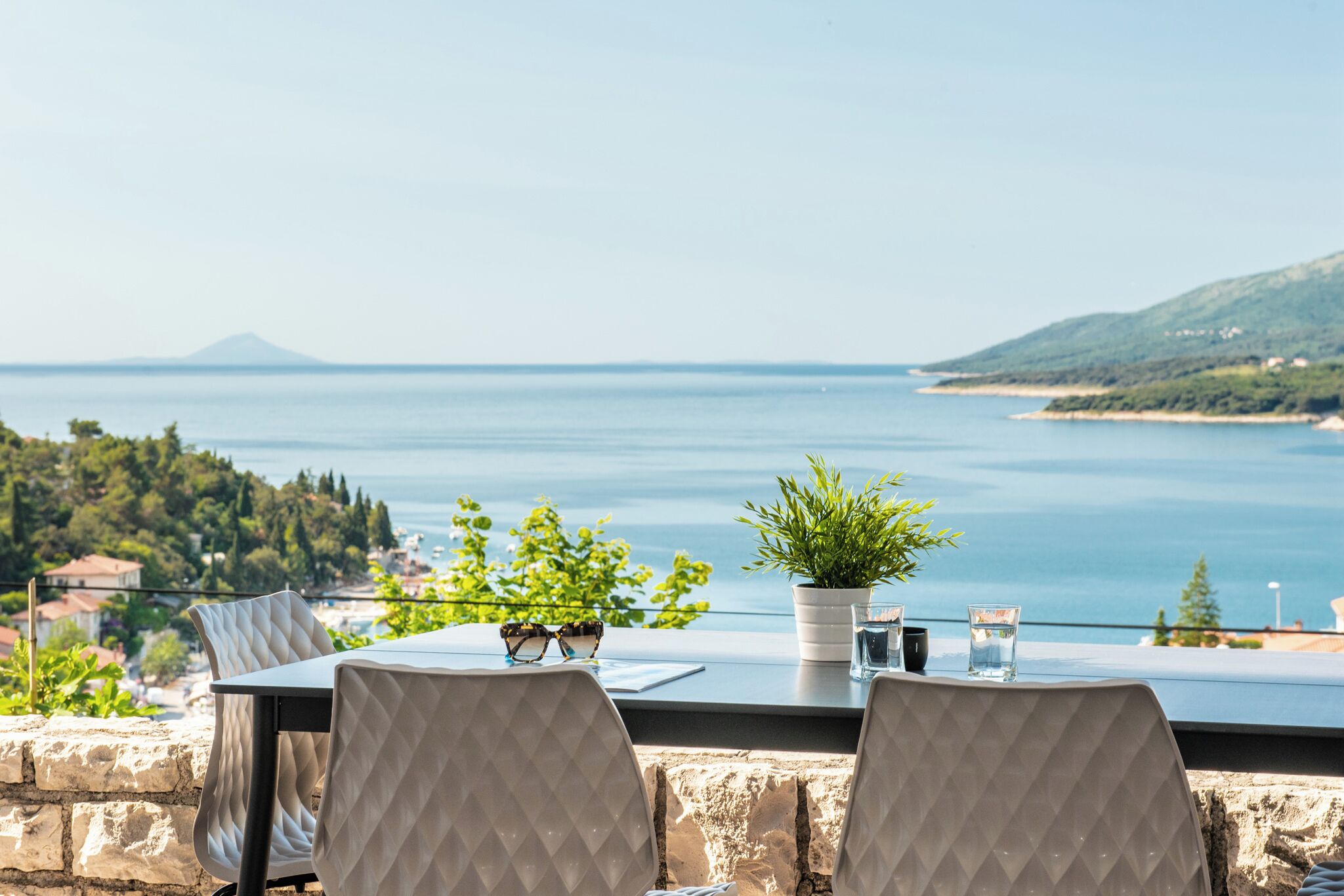 A table with two chairs, a potted plant, a pair of glasses, and a glass of water on a balcony overlooking a body of water and a distant mountain.