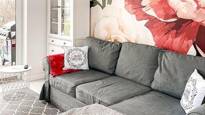 A grey sofa with a red throw blanket and two decorative pillows, a white cabinet with glass doors, and a large floral painting on the wall.