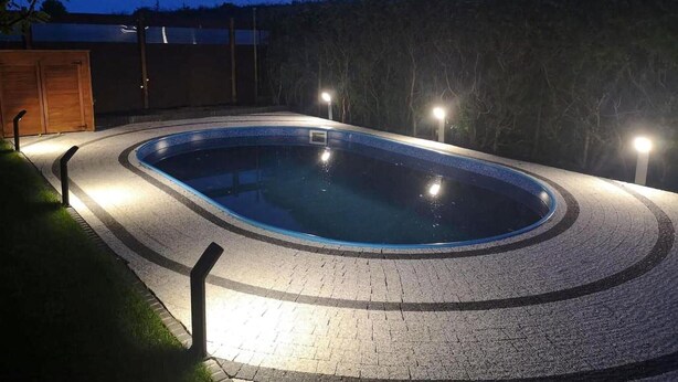A swimming pool with a tiled edge and a brick pathway leading to it, surrounded by a fence and trees.