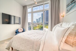 A bedroom with a large bed, a TV mounted on the wall, and a window with a city view.