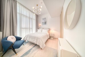 A modern bedroom with a large bed, a blue armchair, a bedside table with a lamp, and a large mirror on the wall. - Bright & Homey Studio Apartment in JLT (Dubai)