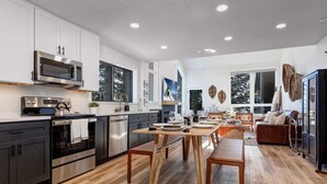 A modern kitchen with stainless steel appliances, a dining area with wooden tables, and a living room with a brown leather sofa. - Remodeled 4BR Chalet | Sleeps 10 | Hot Tub, Pool Access & 5-Min Walk to Lifts (Park City)