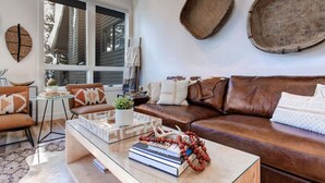 A cozy living room with a brown leather sofa, a glass coffee table with books and decorative items, and a large window providing natural light. - Remodeled 4BR Chalet | Sleeps 10 | Hot Tub, Pool Access & 5-Min Walk to Lifts (Park City)