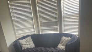 Quarto com sofá azul escuro, estofado com textura acolchoada e botões decorativos, tapete branco e fofo, e poltrona de grãos de espuma azul escuro.