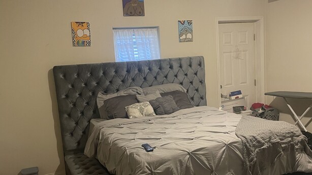 Quarto com uma cama grande, um banquinho pequeno, uma porta e uma janela com cortinas.
