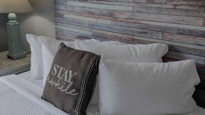Ein Bett mit weißen Kissen und einem grauen Kopfkissen mit der Aufschrift 'STAY awhile', vor einem Holzkopfteil.