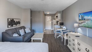 Ein kleines Hotelzimmer mit einer Couch, einem Bett, einem Esstisch, einem Fernseher und einer Küchenzeile.