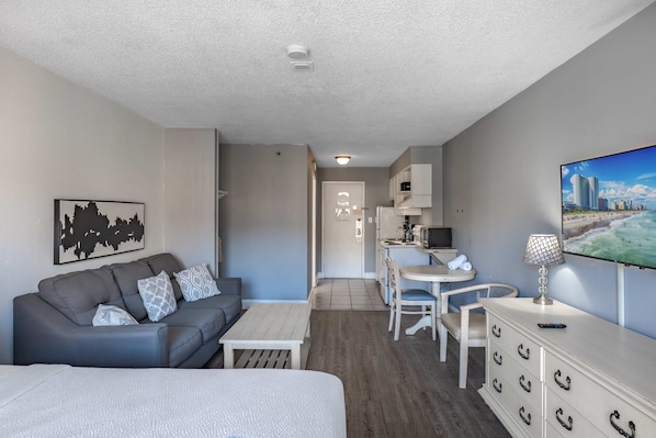 A compact hotel room with a sofa, a bed, a dining table, a TV, and a kitchenette.