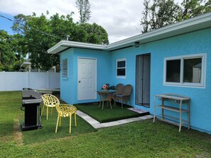 Une maison bleue avec une porte blanche, une fenêtre et un petit patio comprenant une table, des chaises et un barbecue.