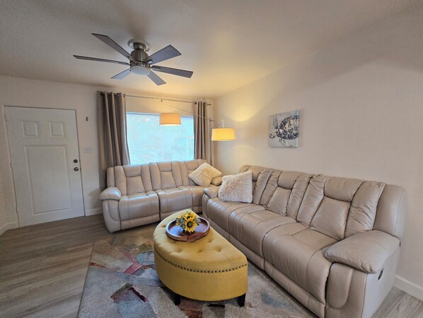 Un salon avec un canapé beige, un pouf jaune, un ventilateur de plafond et une fenêtre avec des stores.