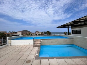 Une piscine sur le toit, avec une eau d’un bleu cristallin, entourée d’une terrasse carrelée, et offrant une vue sur une plage avec des bâtiments en arrière-plan.