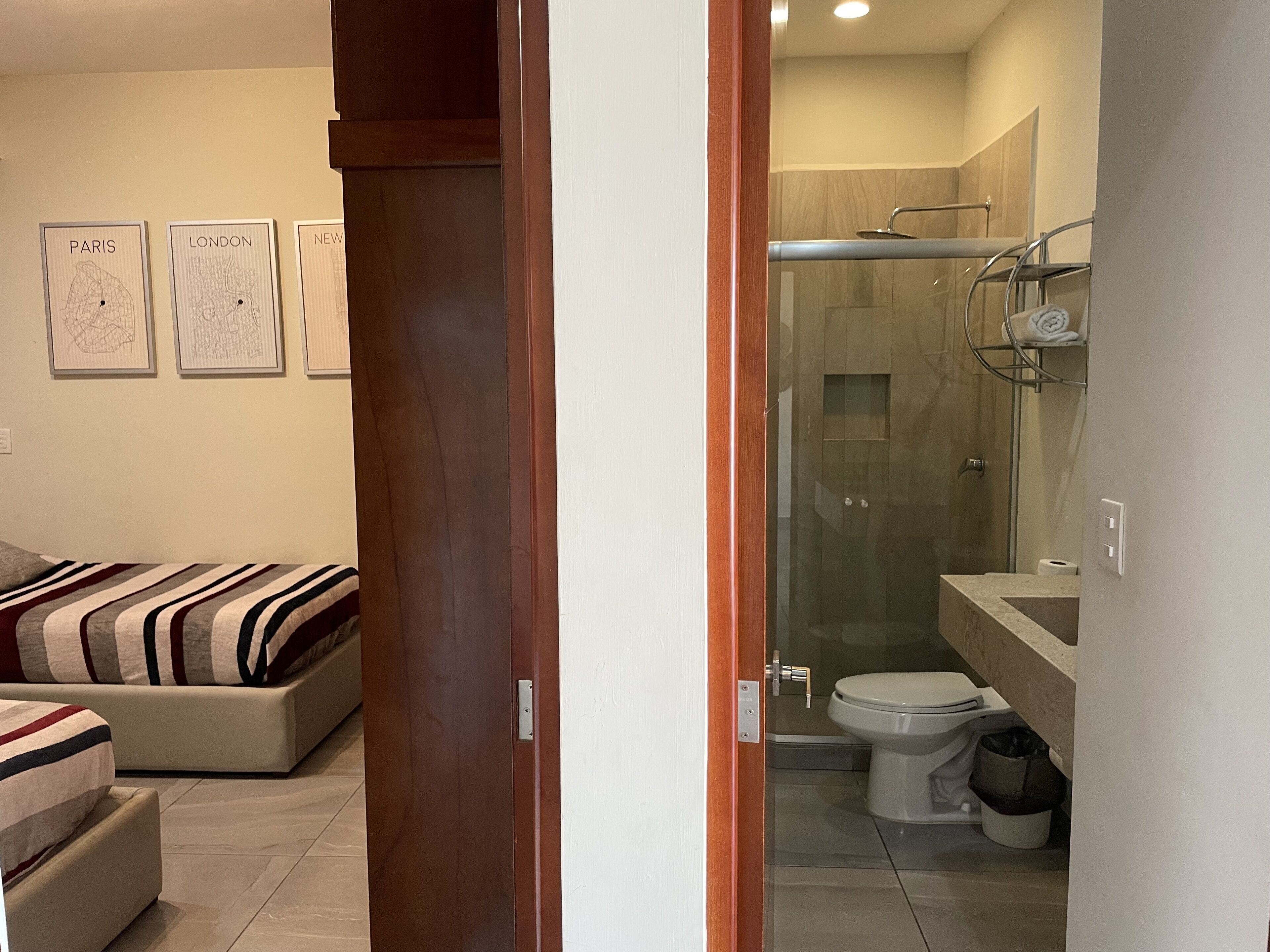 Une salle de bain moderne avec une douche à parois de verre, des toilettes et un lavabo. Des tableaux encadrés ornent le mur, et dans la pièce adjacente, on trouve un lit avec une literie à rayures.