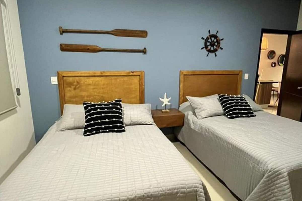 A room with two single beds, each with a wooden headboard, white bedding, and black and white striped pillows. There is a wall-mounted ship's wheel decoration above the beds.