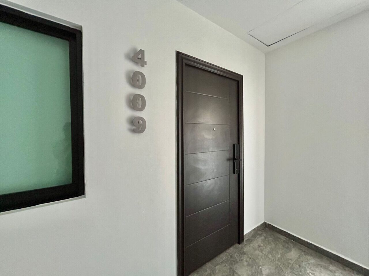 Un pasillo con una puerta oscura, un ventanal con vidrio esmerilado y un letrero con número pegado a la pared.