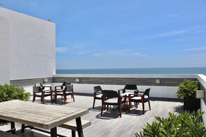 Una terraza en la azotea con mesa y sillas de madera, vistas al océano y plantas en macetas.