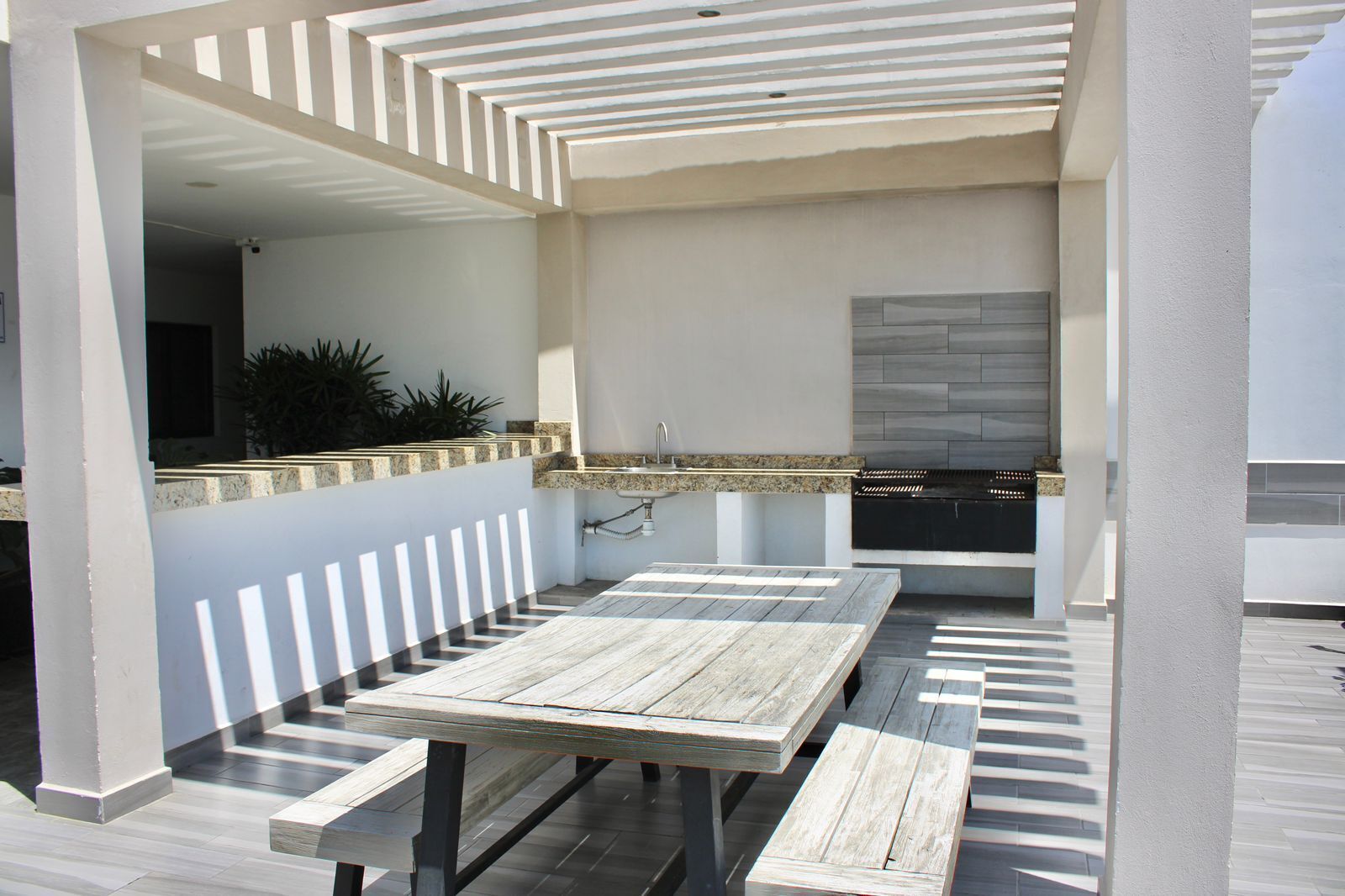 An outdoor patio area with a wooden table and benches, a built-in sink, and a stone backsplash.