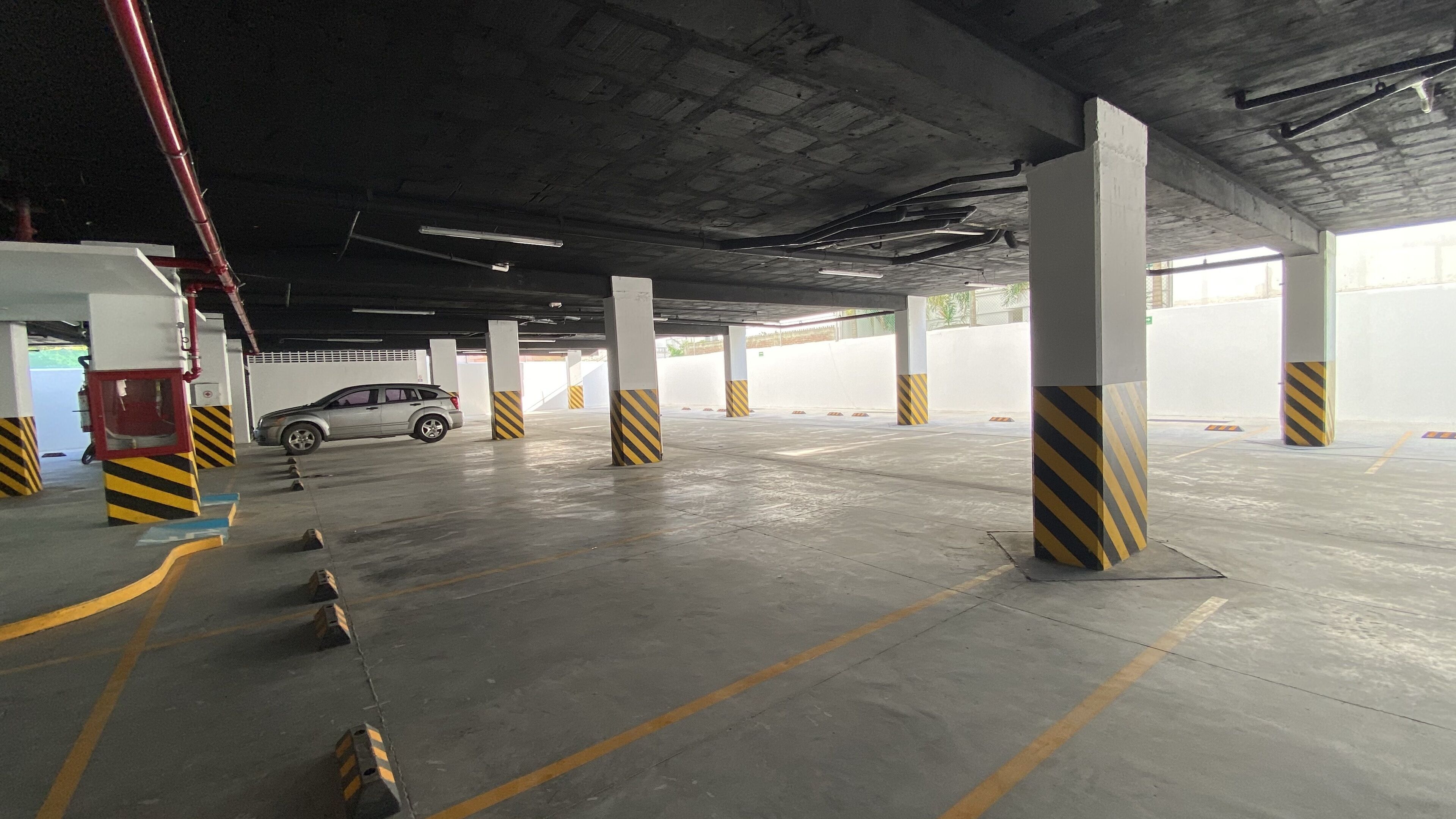 Un estacionamiento interior con pilares de concreto, marcas de seguridad a rayas amarillas y negras, y un coche estacionado a lo lejos.