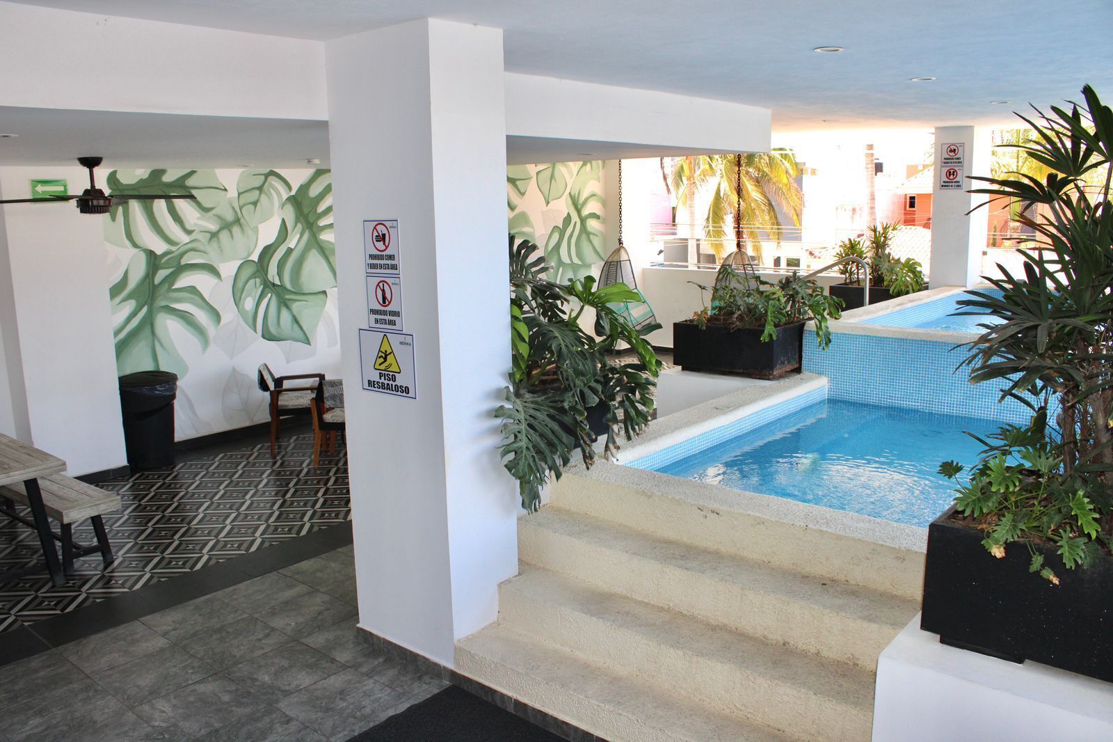 Un'area piscina con scalini che conducono alla vasca, circondata da piante in vaso e un muro decorato con un motivo di foglie tropicali.
