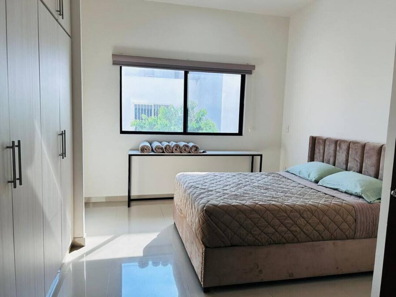 A bedroom with a bed, a window, a bedside table with rolled-up towels, and a wardrobe.