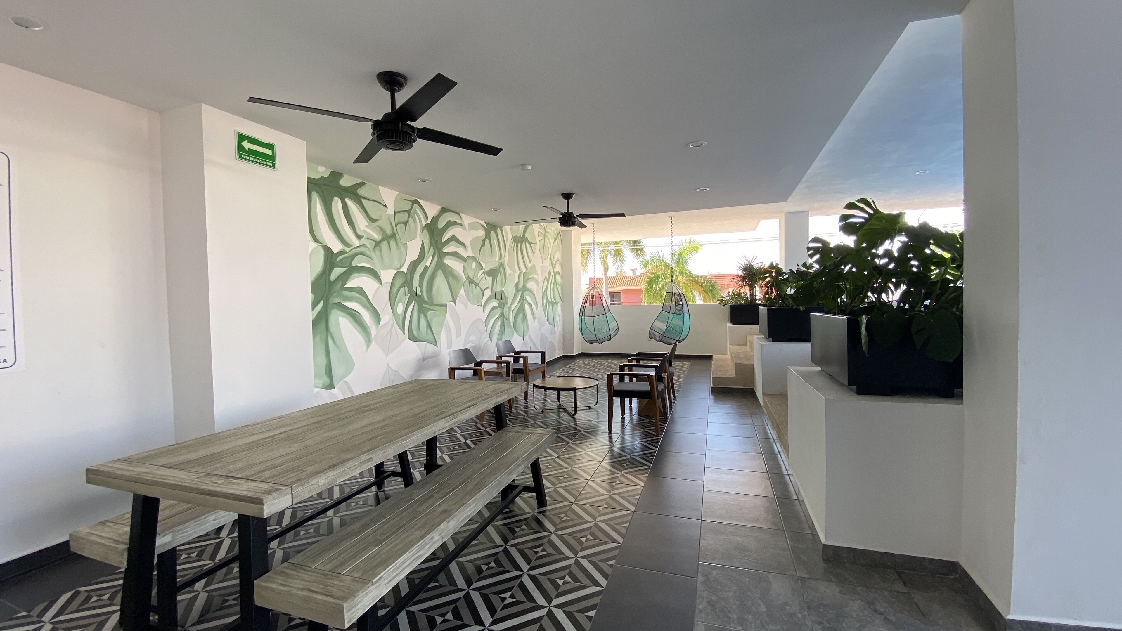 Un espacio interior moderno con un largo banco de madera, ventiladores de techo y una pared con un diseño de hojas.