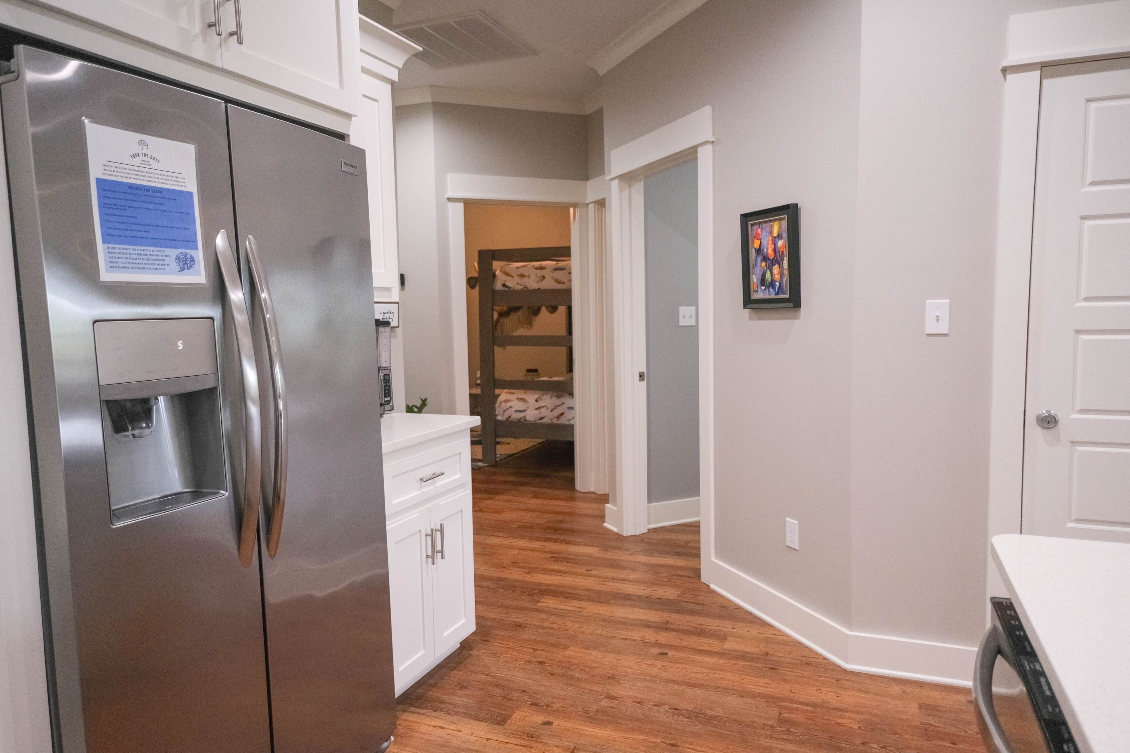 A kitchen with a stainless steel refrigerator, wooden flooring, white cabinetry, and a hallway leading to a bedroom with a bunk bed.