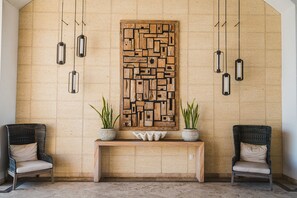 A lobby with a wooden wall art piece, two wicker chairs, a bench, and hanging pendant lights.