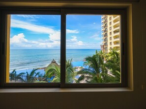 A view through a window of a hotel with a pool and palm trees, overlooking the ocean and a building with balconies.