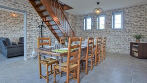 A dining area with a wooden table set for six, surrounded by wooden chairs. A staircase with a wooden railing leads to an upper level. The walls are made of stone, and there are two windows providing natural light.