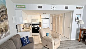 A living room with a beige sofa and a recliner, a kitchen with white cabinets and appliances, and a hallway leading to a door. - What if I told you…You CAN afford Paradise! Make the Call today! 🤙 (Gulf Shores)