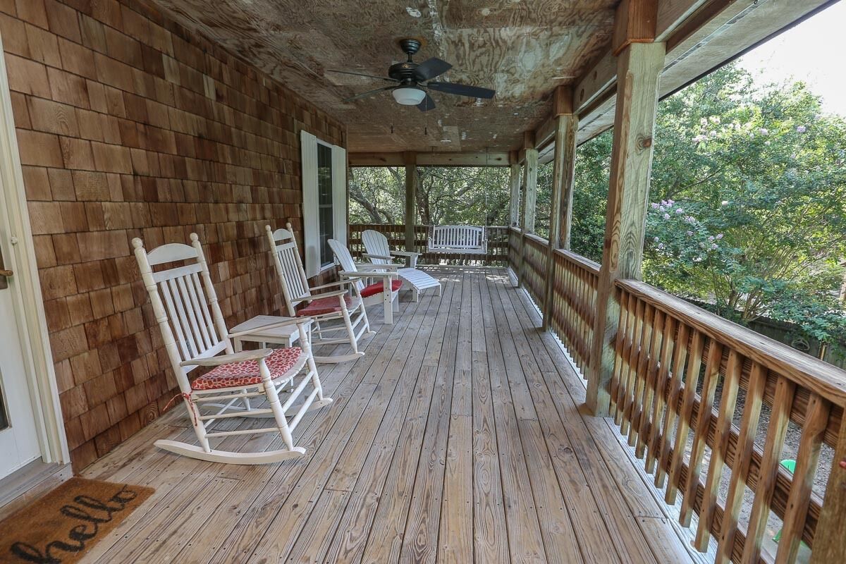A wooden porch with rocking chairs, a ceiling fan, and a view of trees.