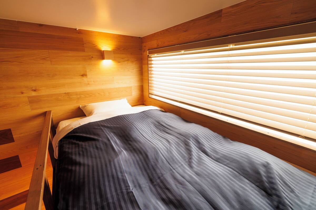 A small, cozy bedroom with a wooden headboard, a single bed with striped bedding, and a window with blinds.