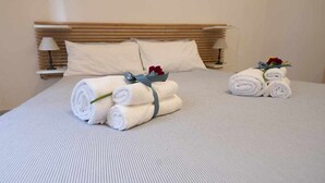 Un lit bien fait, avec des serviettes blanches pliées en rouleau et un petit bouquet de fleurs, une tête de lit en bois et une table de chevet avec une lampe.