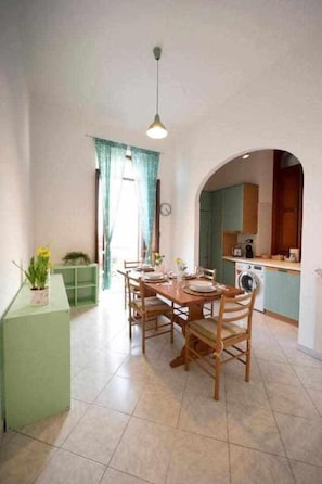 Un coin repas avec une table et des chaises en bois, un buffet vert agrémenté d’une plante, et une cuisine visible à travers une arche.