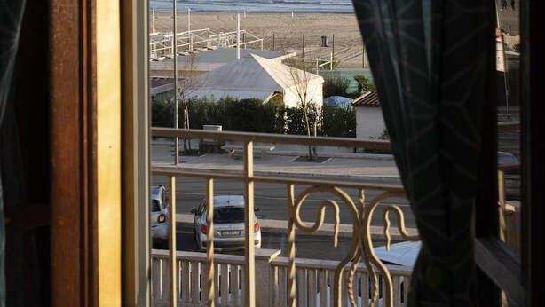Une vue depuis une fenêtre, à travers des rideaux à motifs, donnant sur une plage avec des bâtiments et une rue.