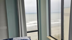 A bedroom with a large window overlooking a beach, a bed with a blue and white quilt, and a curtain rod. - Beautiful Oceanfront Top-Floor Condo, Pool, Beach View, (North Myrtle Beach)