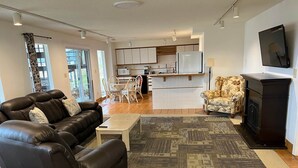 A modern living room with a leather sofa, a coffee table, and a patterned rug. There is a kitchen area with white cabinets and a flat-screen TV mounted on the wall. - Lazy O Resort - 7 bedroom 3 bathroom house. (Camdenton)