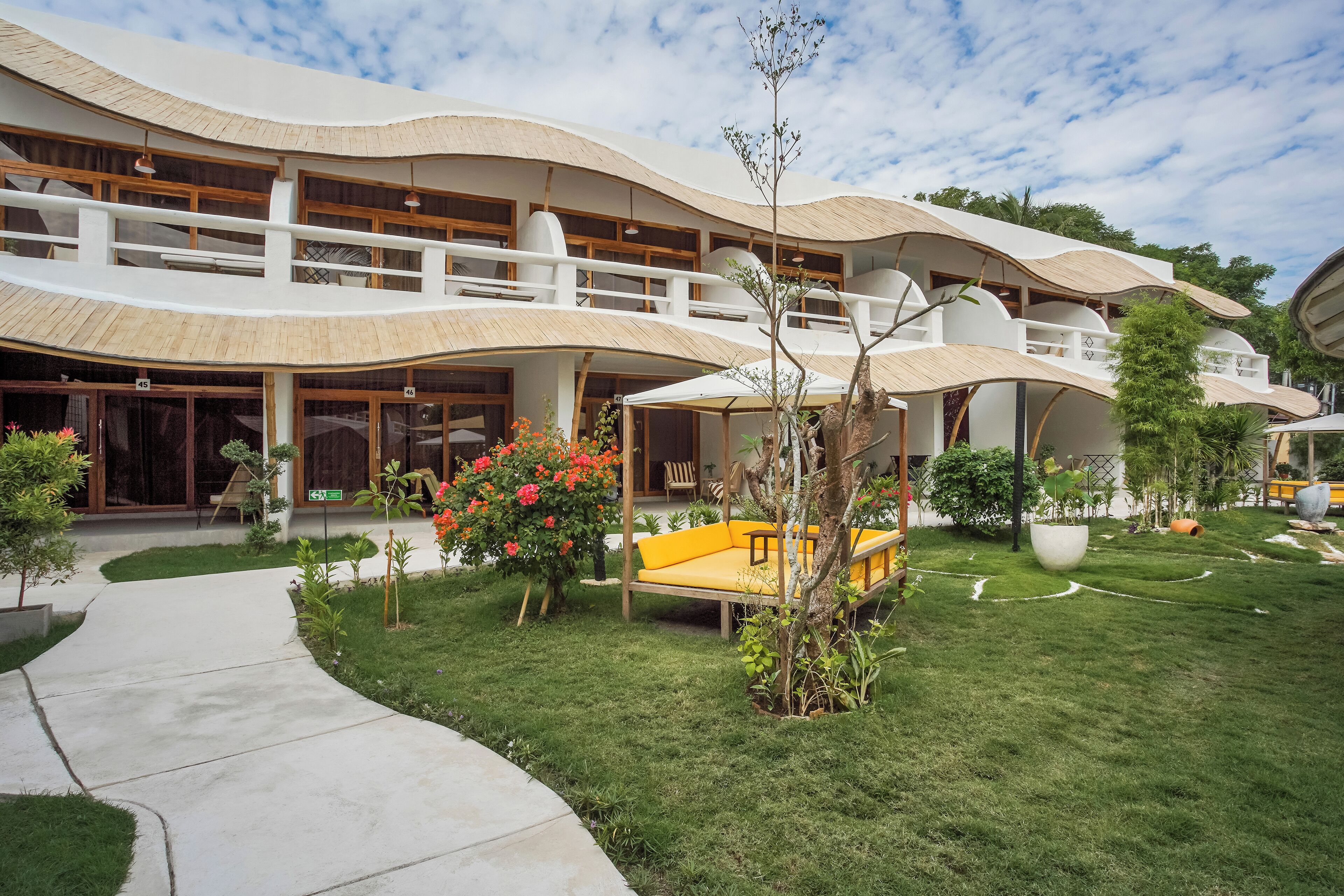 Il s’agit d’un bâtiment moderne doté d’un toit incurvé et de nombreux balcons. Un chemin mène à l’entrée, et un banc jaune est visible dans le jardin.