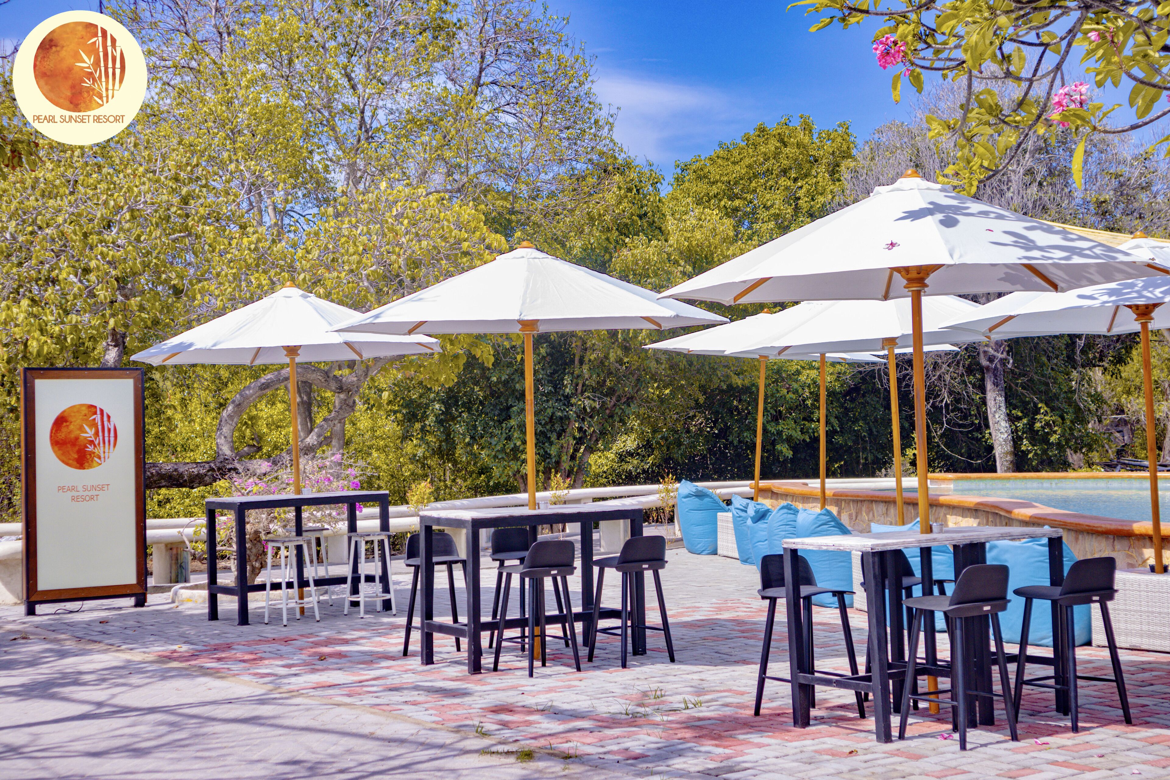 An outdoor seating area with tables and chairs, umbrellas, and a pool in the background.
