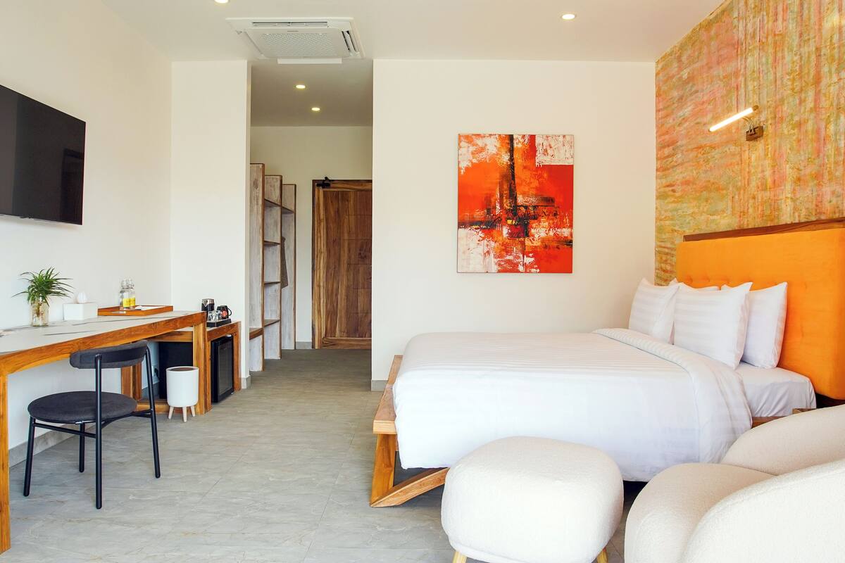A modern hotel room with a bed, a desk, and a chair. There is a painting on the wall and a television mounted on the wall.