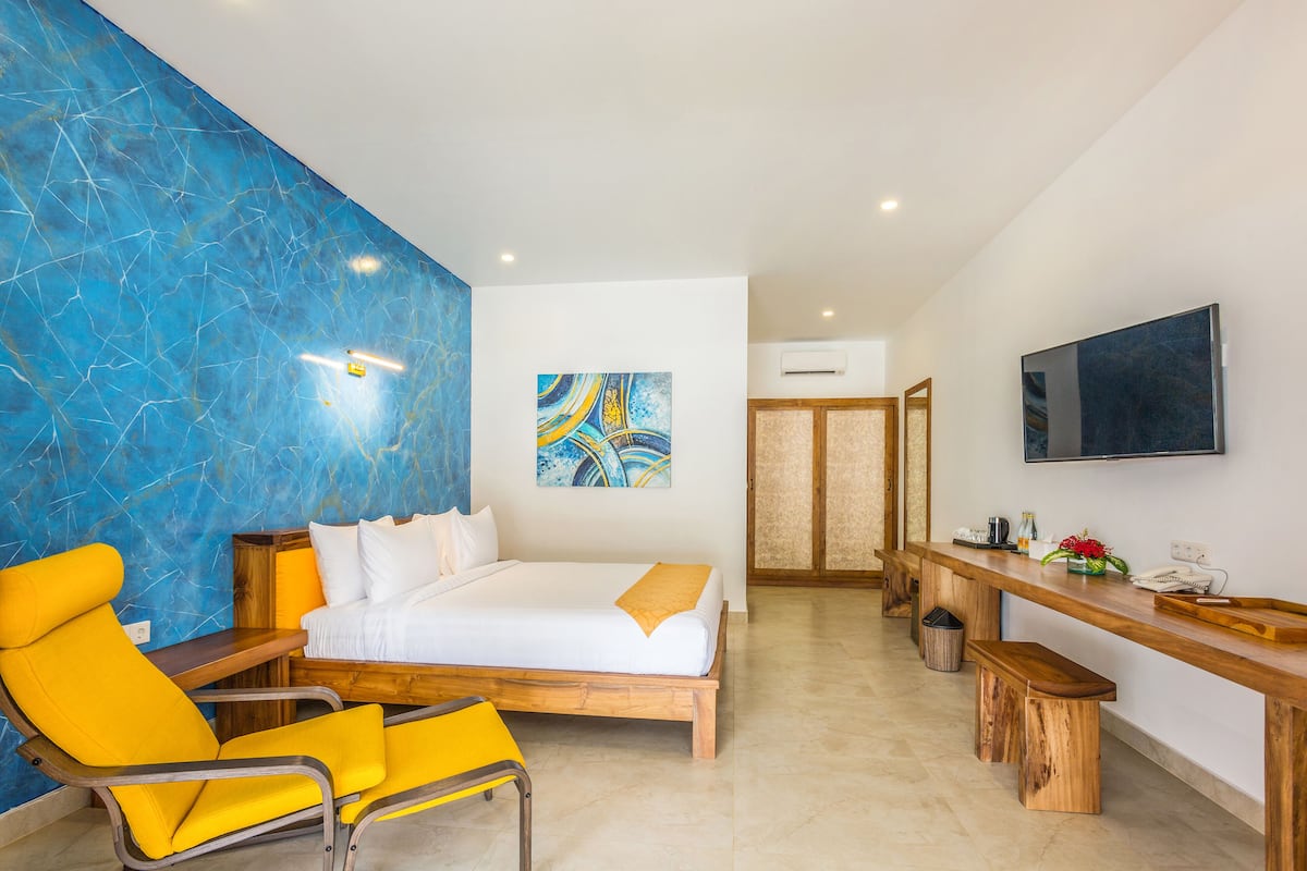 A hotel room with a bed, a wooden desk, a yellow armchair, a blue marble wall, and a mounted TV.