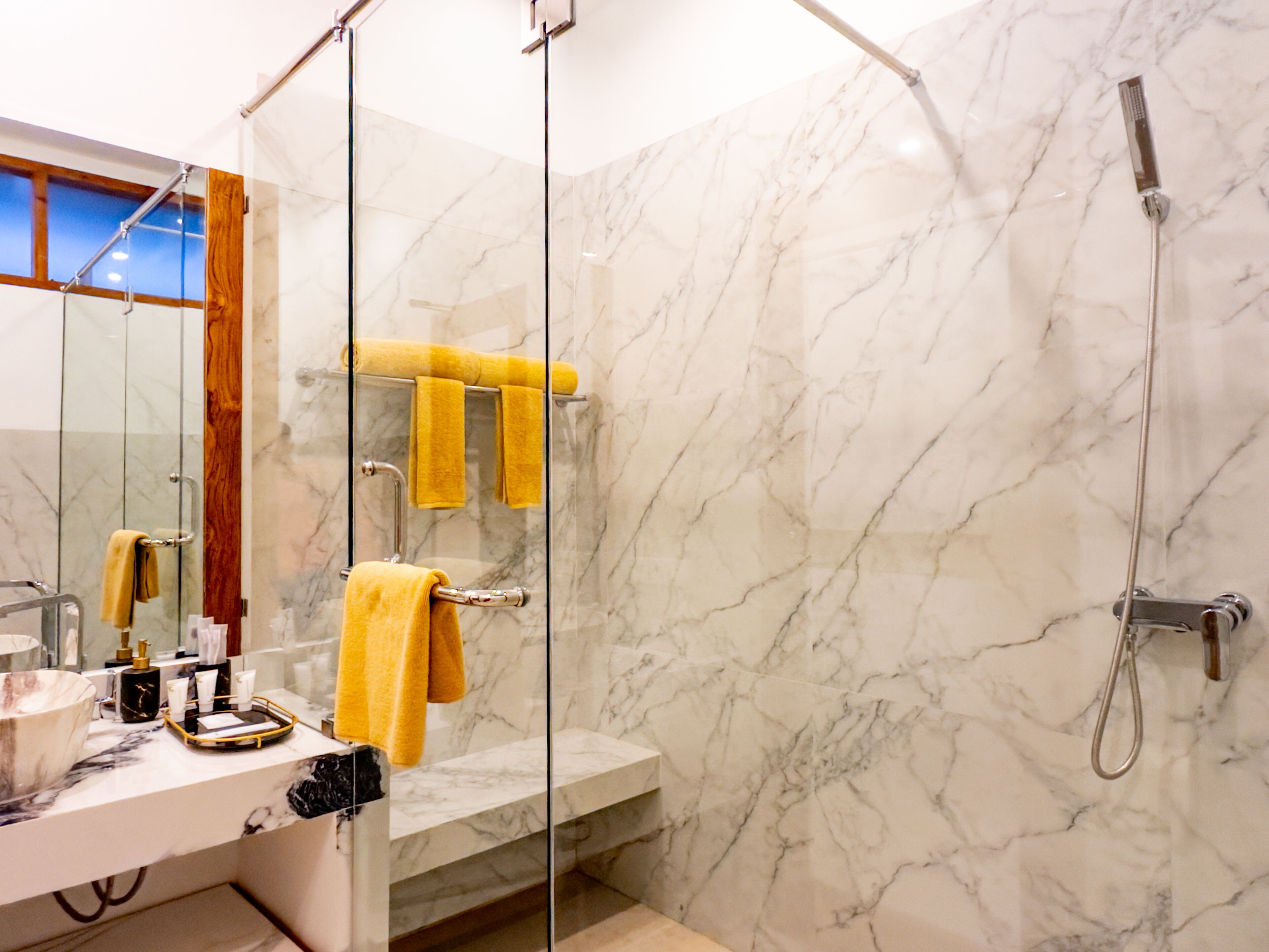 Une salle de bain avec un mur en marbre, une douche en verre, un lavabo avec des articles de toilette et un miroir avec un cadre en bois.