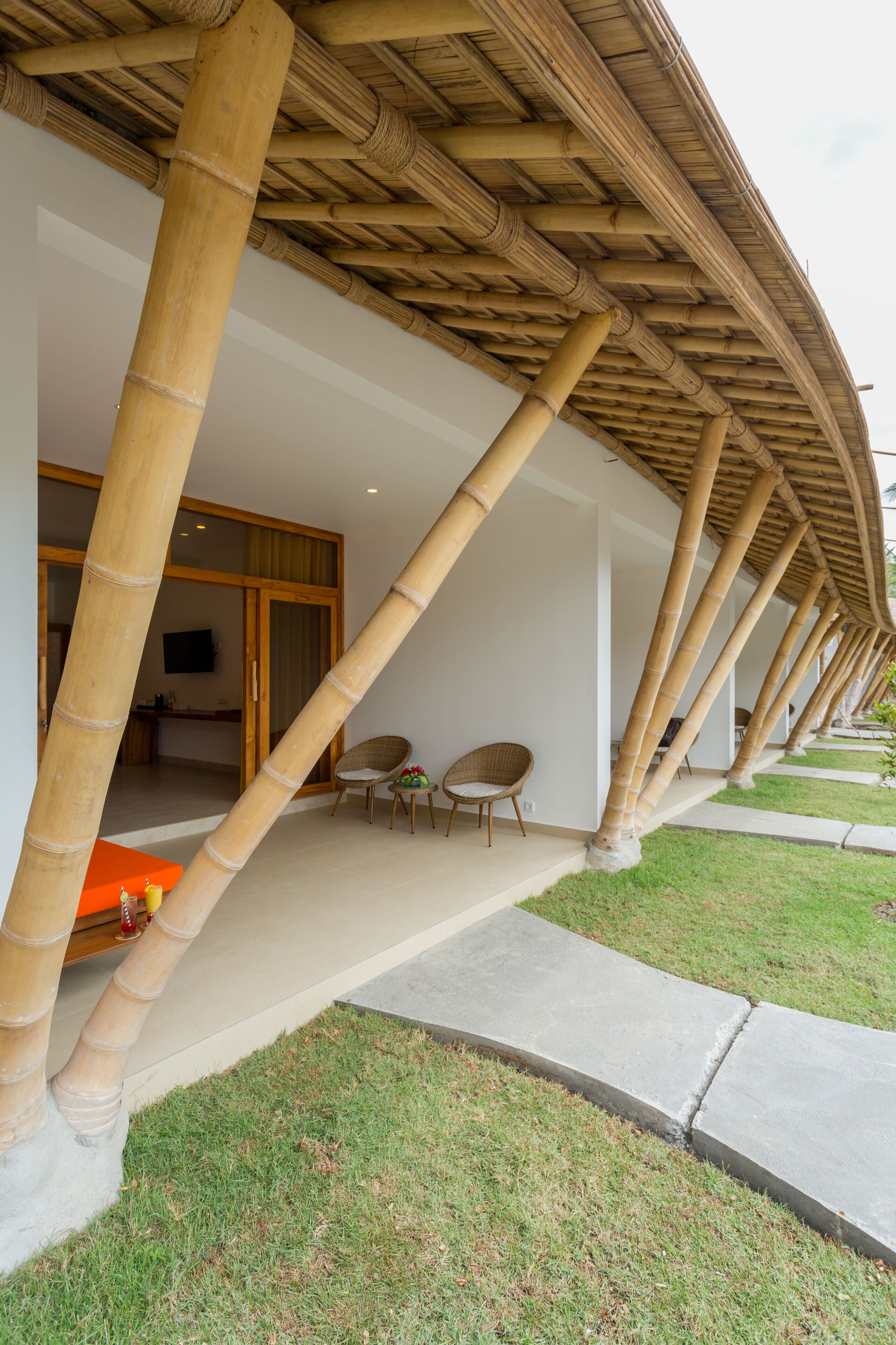 Un espace extérieur abrité, doté de piliers en bambou, un coin salon avec des chaises, et un chemin menant à l’entrée d’un bâtiment.