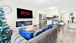 Una sala de estar moderna con un sofá gris, un televisor de pantalla plana, un árbol de Navidad y una cocina al fondo.