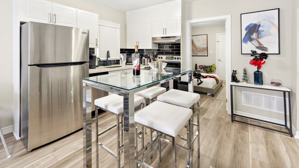 Una cocina moderna con electrodomésticos de acero inoxidable, una isla de vidrio con taburetes y un comedor con mesa y sillas blancas.