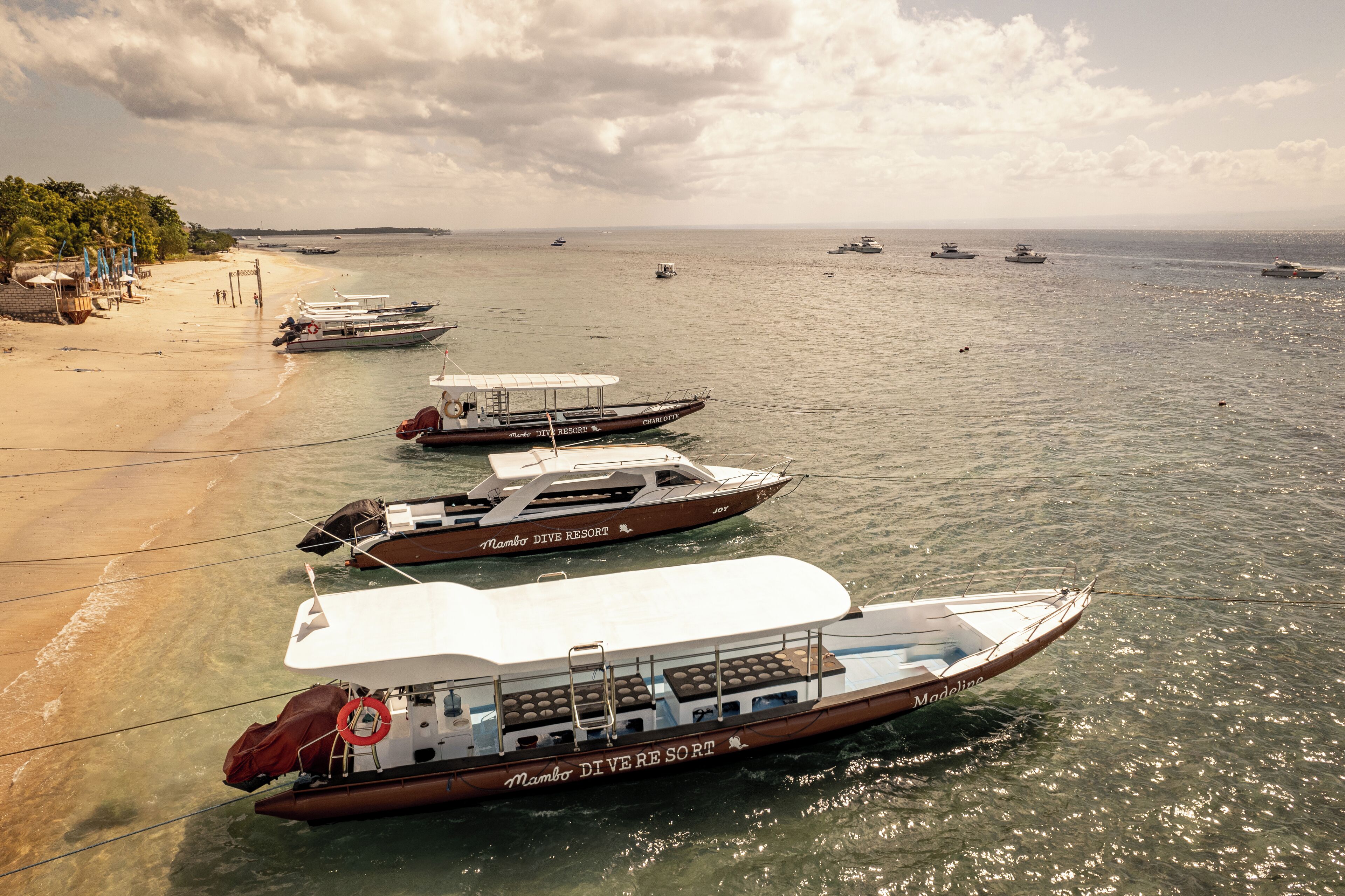 Una fila de barcos anclados en una playa con agua cristalina y un cielo nublado.
