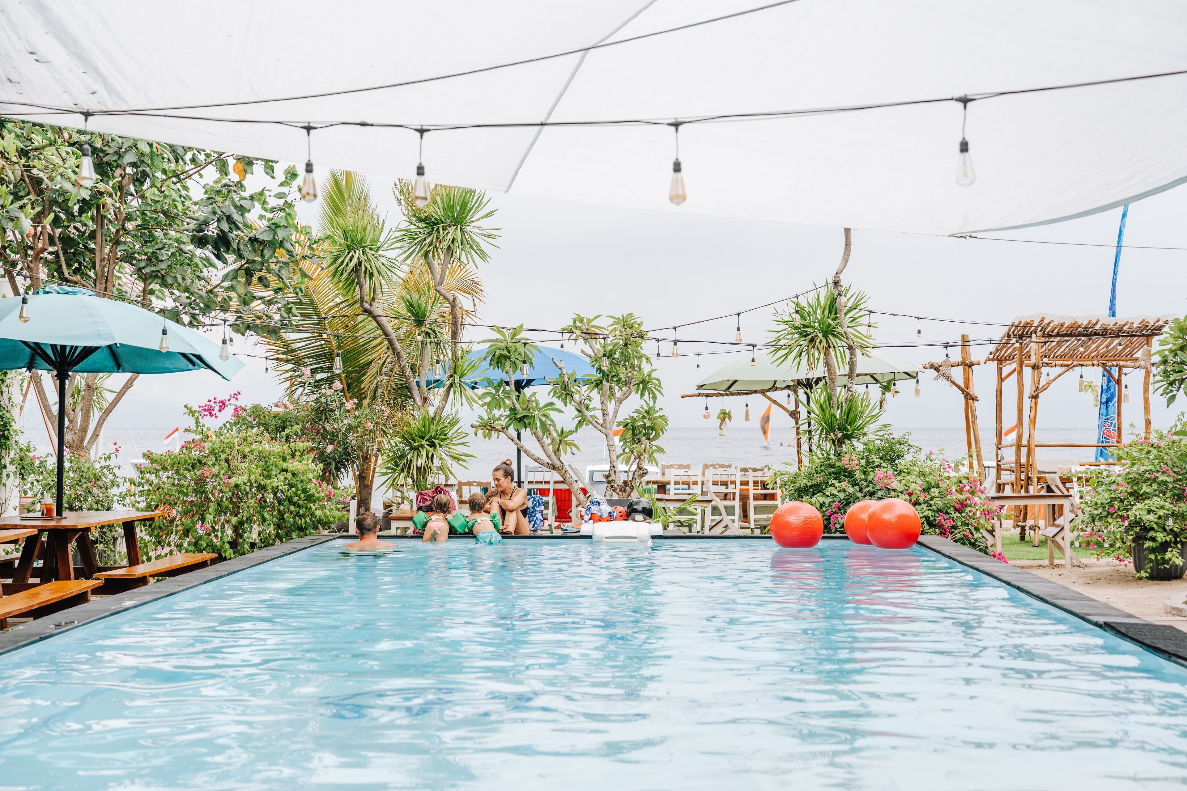An outdoor swimming pool with wooden benches and umbrellas, surrounded by tropical plants and a wooden pergola.