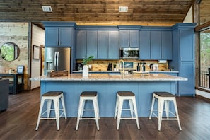 A modern kitchen with blue cabinets, a stainless steel refrigerator, and a granite countertop. There are three white stools and a large window providing natural light.