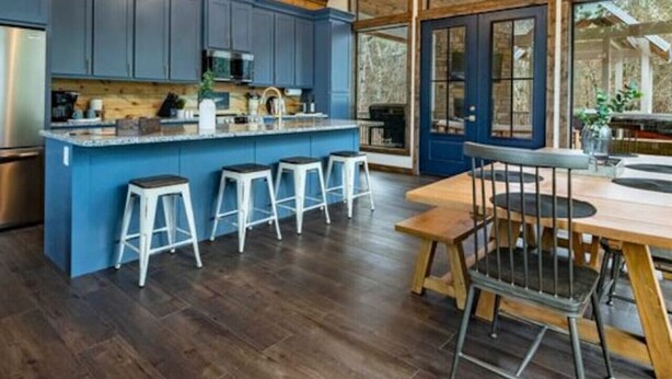 A modern kitchen with blue cabinets, a central island with stools, and a large window overlooking a natural landscape. - Luxury Cabin! Hot tub, Pet Friendly, Foosball! (Broken Bow)
